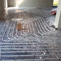 Rozłożona instalacja ogrzewania podłogowego z rurkami na folii odblaskowej, widok z góry, w tle surowe ściany i okno balkonowe, na instalacji leży karton z numerem 3.