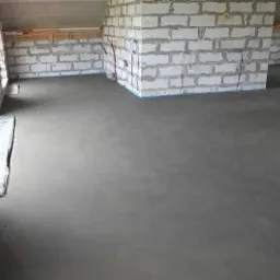 Świeżo wylana, gładka, szara posadzka w surowym wnętrzu z białych bloczków, zabezpieczona folią przy oknie.