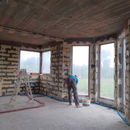 PUH Michał Zulczyk - Wnętrze w trakcie prac wykończeniowych: mur z widocznymi spoinami, częściowo pokryty siatką i zaprawą, instalacje elektryczne i hydrauliczne na ścianach, pracownik w kombinezonie wyrównuje ścianę...