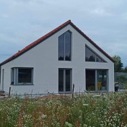 PUH Michał Zulczyk - Nowoczesny dom jednorodzinny z białymi ścianami zewnętrznymi i czerwoną dachówką, widoczny z perspektywy łąki z kwitnącymi kwiatami, w tle inne domy i drzewa.