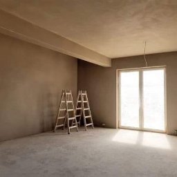 PUH Michał Zulczyk - Surowe wnętrze po nałożeniu tynku, z dwoma drewnianymi drabinami opartymi o ścianę, widok na okno z intensywnym światłem wpadającym do środka, przygotowane do dalszych prac wykończeniowych.