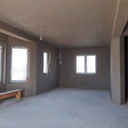 PUH Michał Zulczyk - Wnętrze surowego pomieszczenia po nałożeniu tynku, z widocznymi trzema oknami zabezpieczonymi folią, drewnianym stołkiem pod oknem oraz wystającymi przewodami elektrycznymi z sufitu.