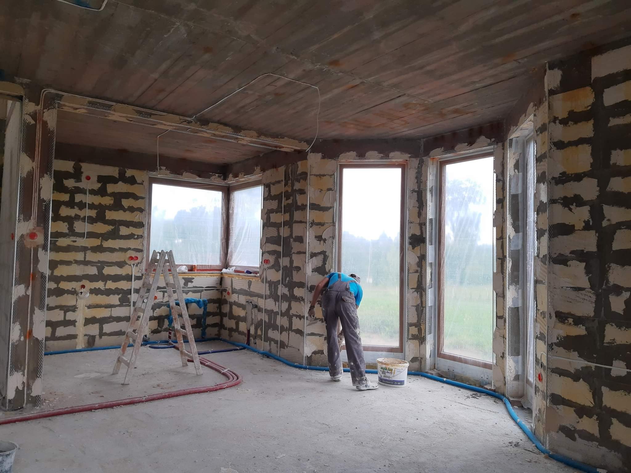 Wnętrze w trakcie prac wykończeniowych: mur z widocznymi spoinami, częściowo pokryty siatką i zaprawą, instalacje elektryczne i hydrauliczne na ścianach, pracownik w kombinezonie wyrównuje ścianę...
