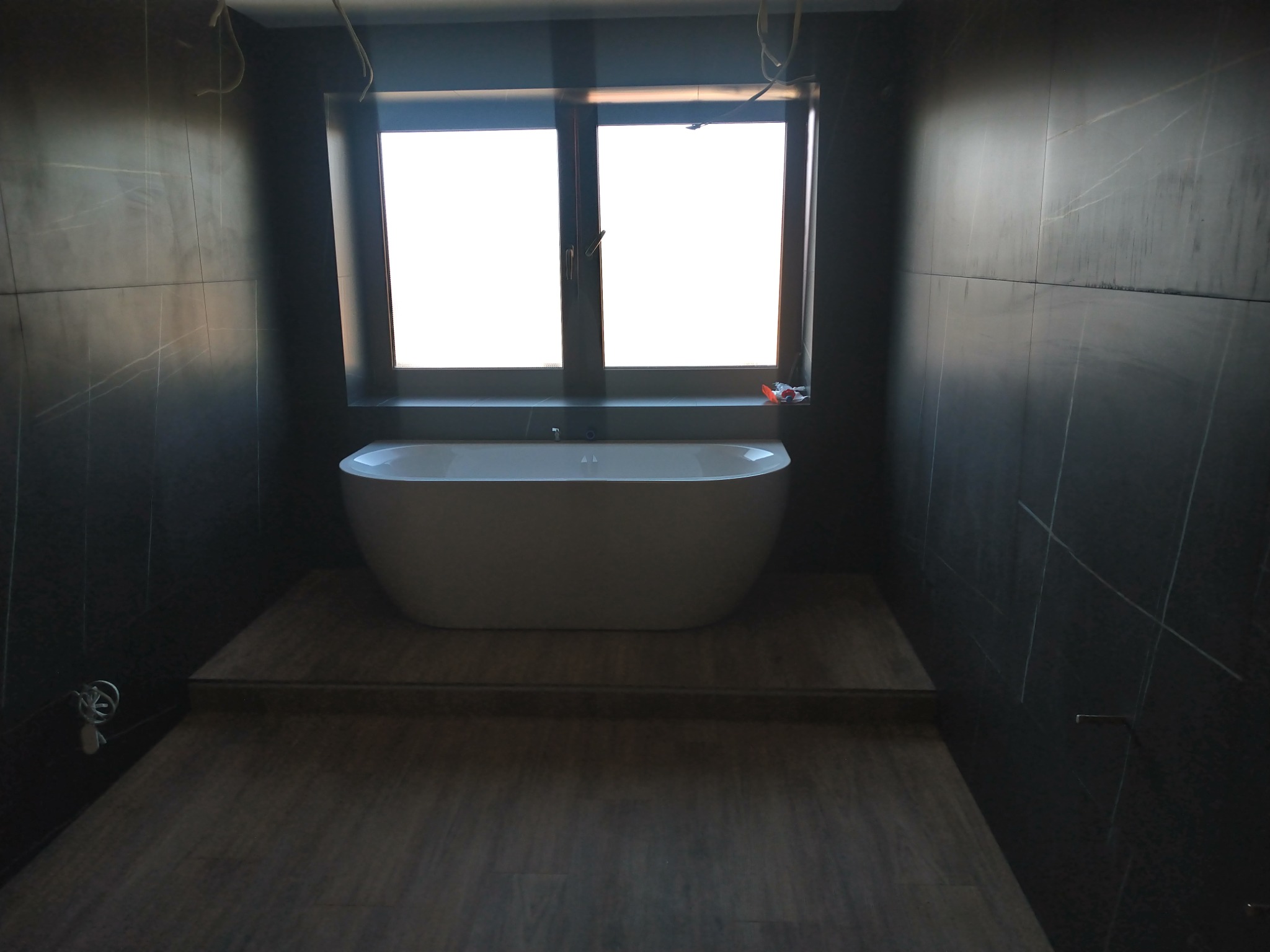 Nowoczesna łazienka w trakcie wykańczania, z wolnostojącą białą wanną na podwyższeniu z drewnopodobnych paneli i ciemnymi płytkami na ścianach, widoczne przewody elektryczne i fragment okna.