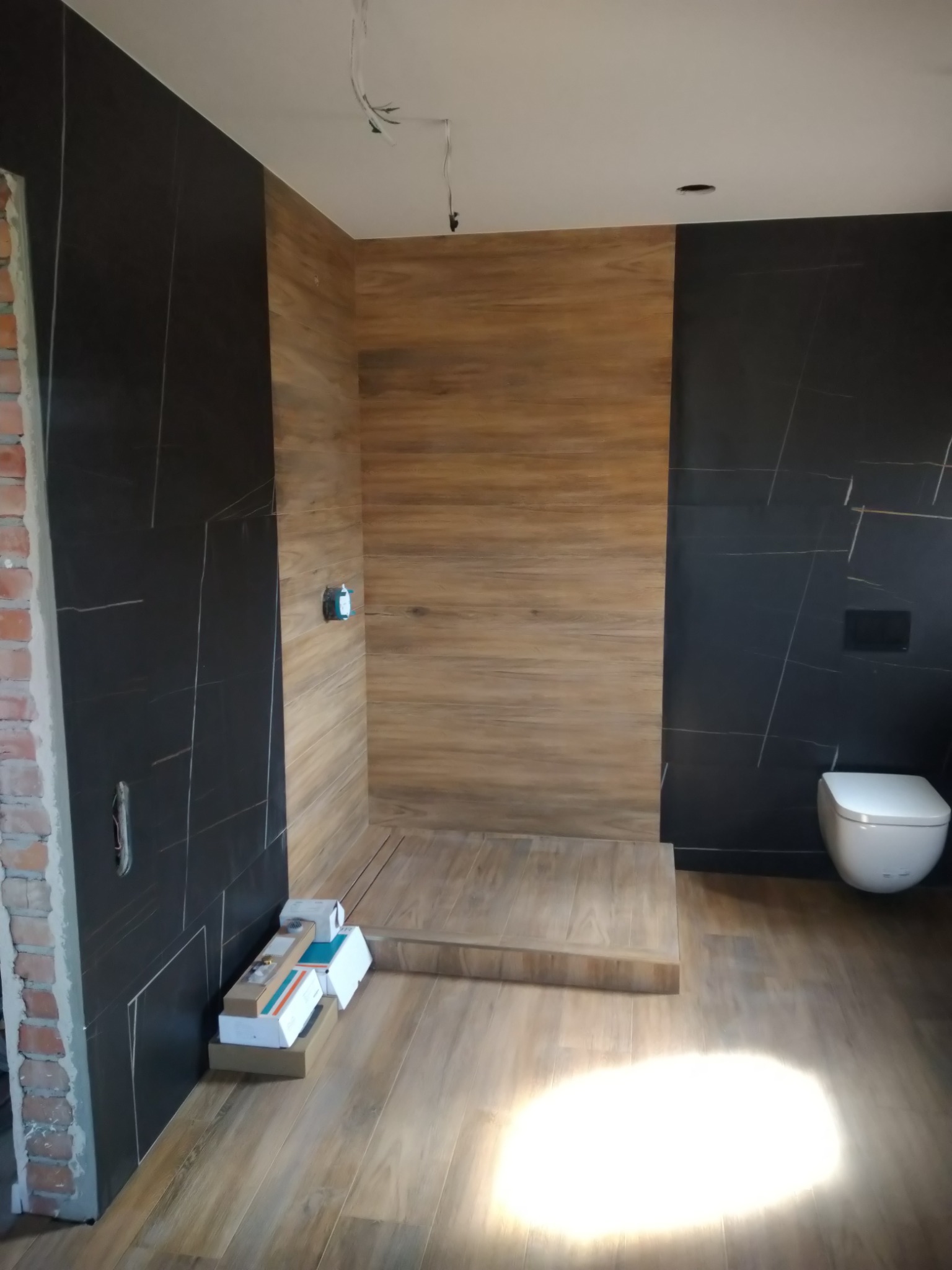 Wnętrze łazienki w trakcie prac wykończeniowych: czarne płytki z białymi żyłkami na ścianach, imitacja drewna na podłodze i w strefie prysznicowej, częściowo wykończony mur przy wejściu, kartony...