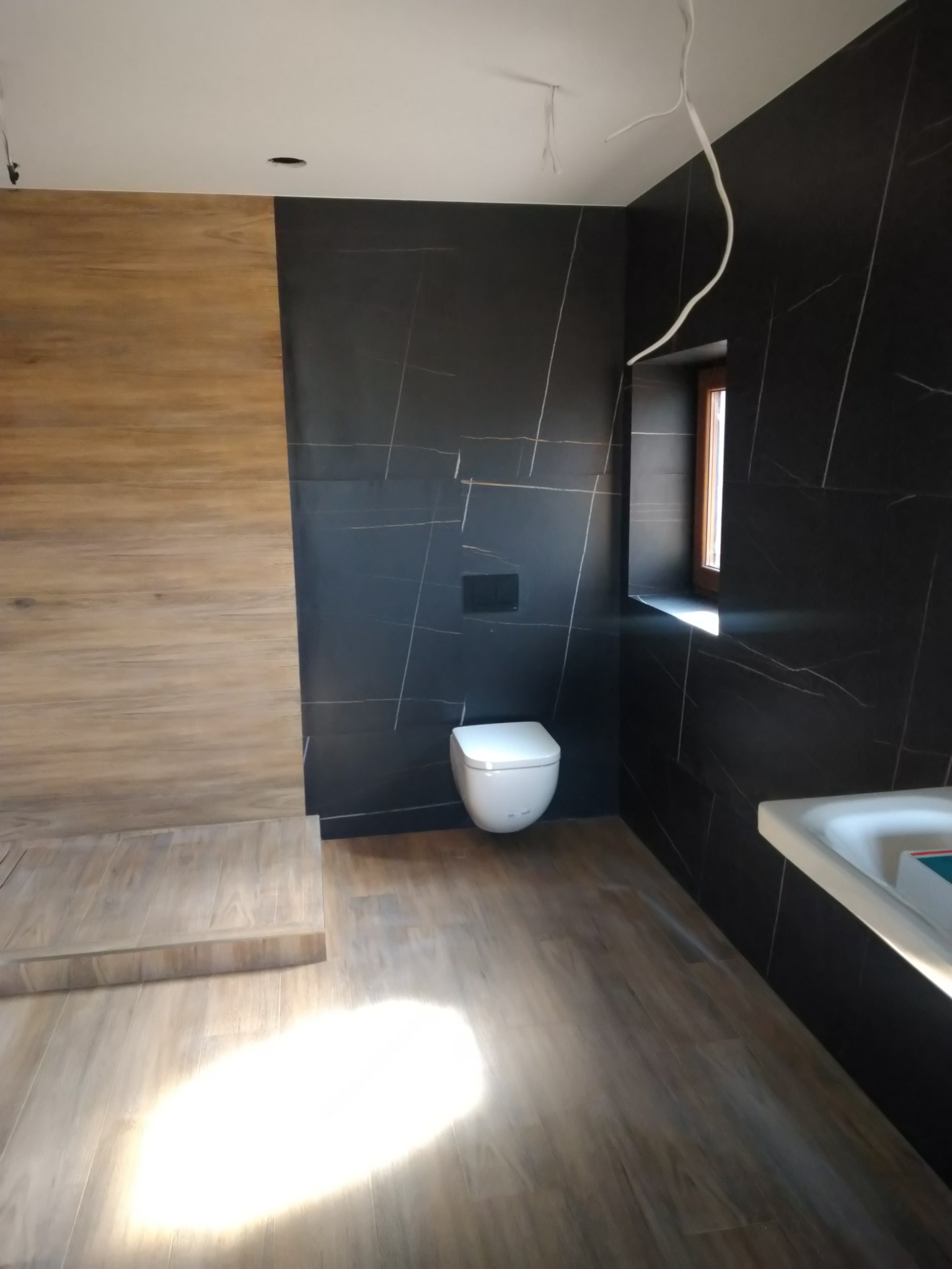 Nowoczesna łazienka z czarnymi płytkami imitującymi marmur z białymi żyłami, drewnopodobną podłogą i ścianą, oraz wiszącą toaletą. Widoczne niedokończone podłączenie elektryczne.