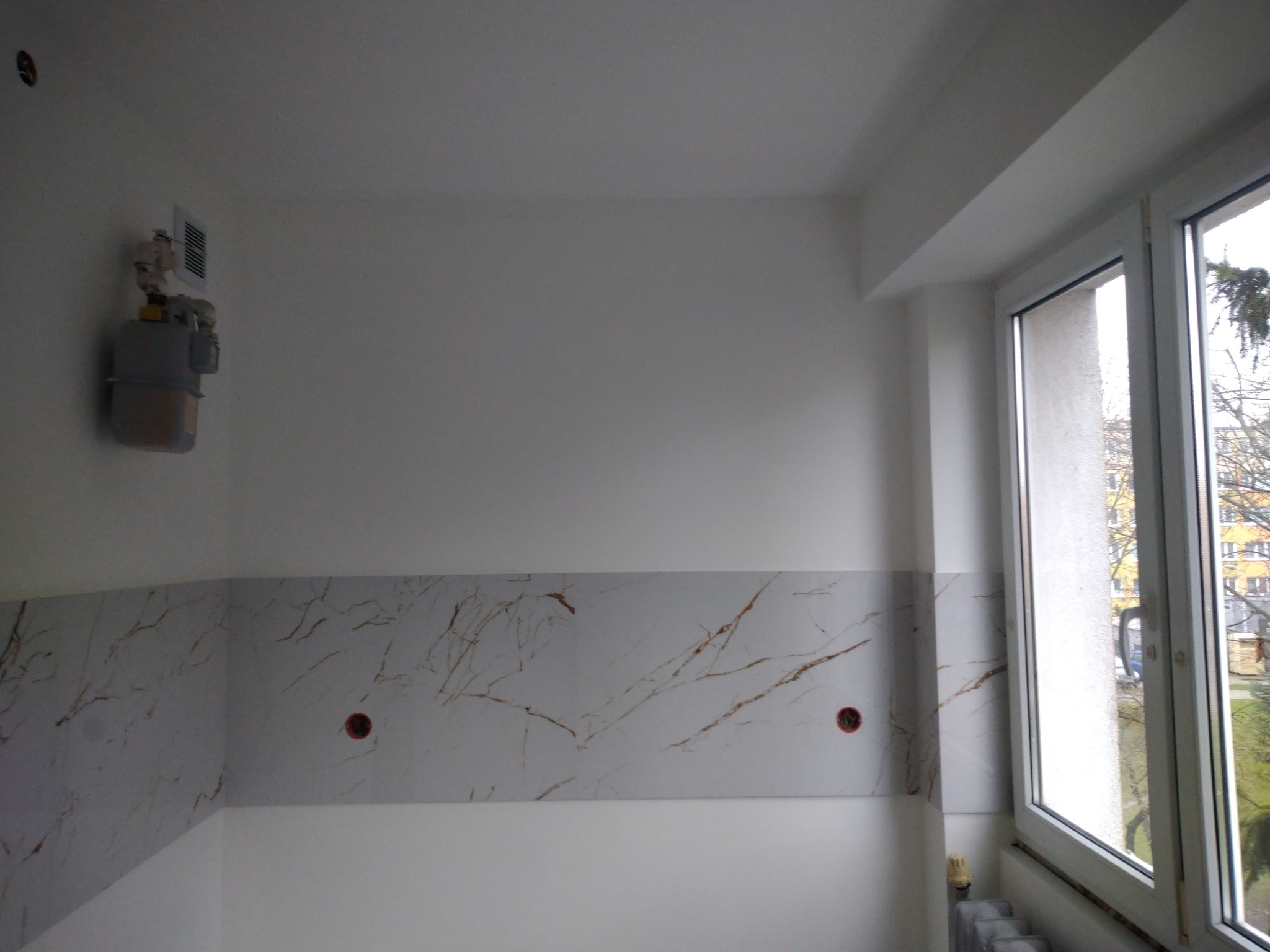 Fragment ściany z panelami imitującymi marmur z widocznymi gniazdkami elektrycznymi, obok okno z widokiem na drzewa i budynki, na ścianie licznik gazu.