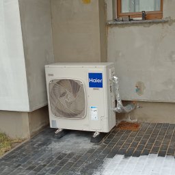 Paweł Hyża - Zielony Amper - Zewnętrzna jednostka klimatyzacji Haier 8kW zamontowana przy ścianie budynku, z widocznymi rurami w izolacji aluminiowej i fragmentem okna na tle surowego tynku.