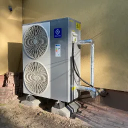 Szara jednostka zewnętrzna z dwoma wentylatorami marki Nulite zamontowana na betonowych podstawach przy żółtej ścianie budynku, z izolowanymi rurami i stertą cegieł obok.