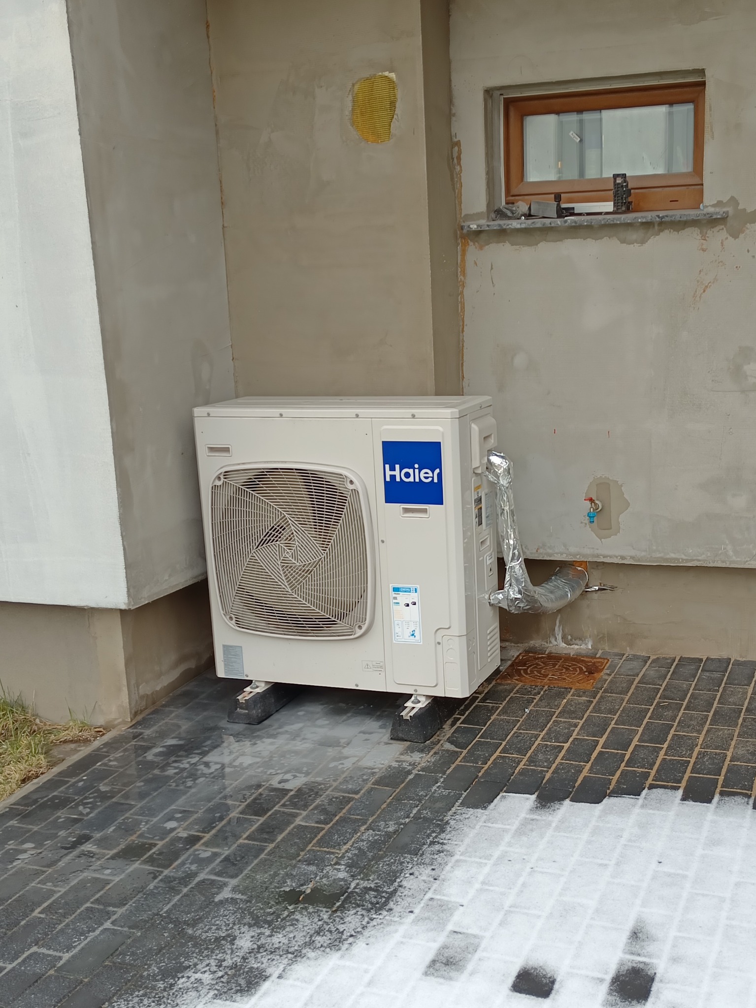Zewnętrzna jednostka klimatyzacji Haier 8kW zamontowana przy ścianie budynku, z widocznymi rurami w izolacji aluminiowej i fragmentem okna na tle surowego tynku.