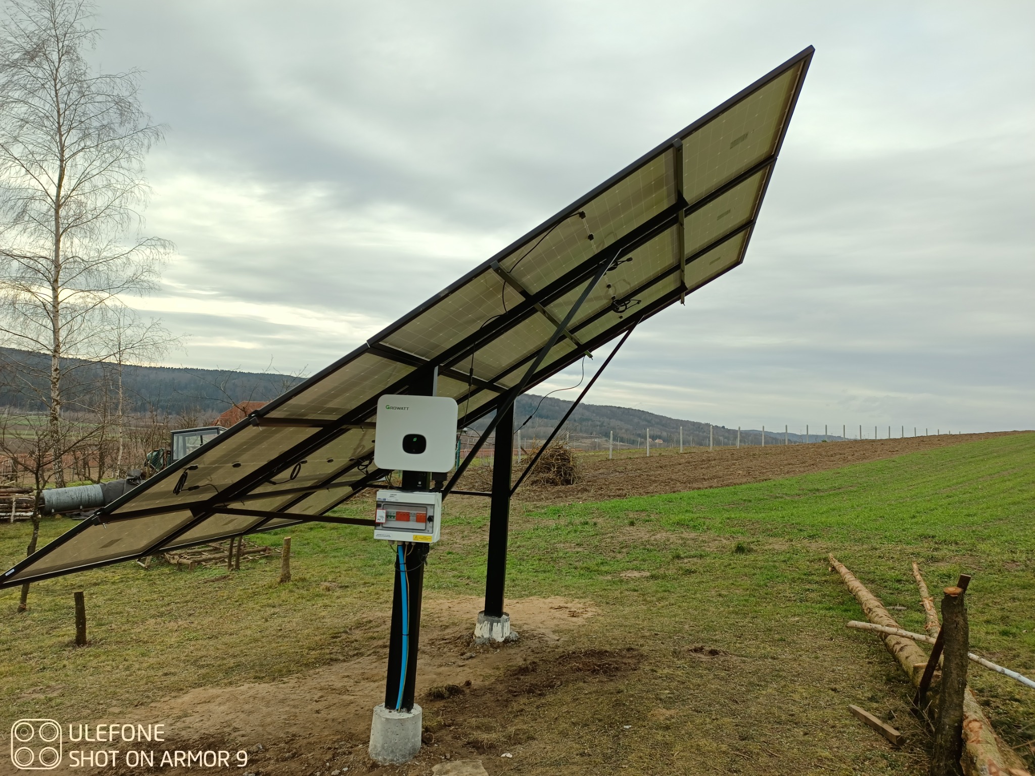 Instalacja fotowoltaiczna DAH Solar o mocy 3,3kWp z inwerterem Growatt na tle pola i pochmurnego nieba, widoczna skrzynka elektryczna z bezpiecznikami.