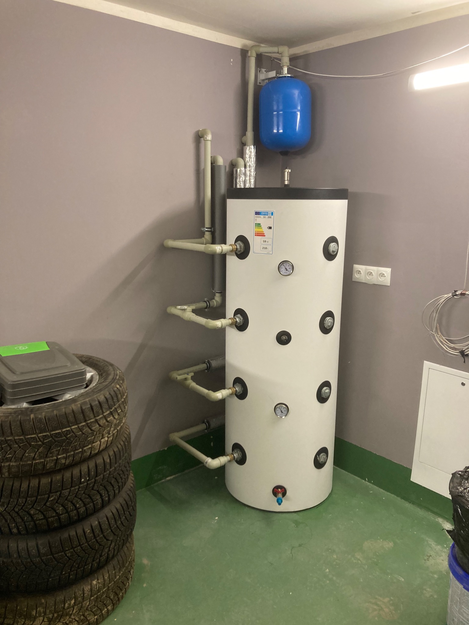 Biały, cylindryczny zbiornik z manometrami i niebieskim zbiornikiem wyrównawczym w szarej kotłowni, obok sterta opon samochodowych i skrzynka narzędziowa.