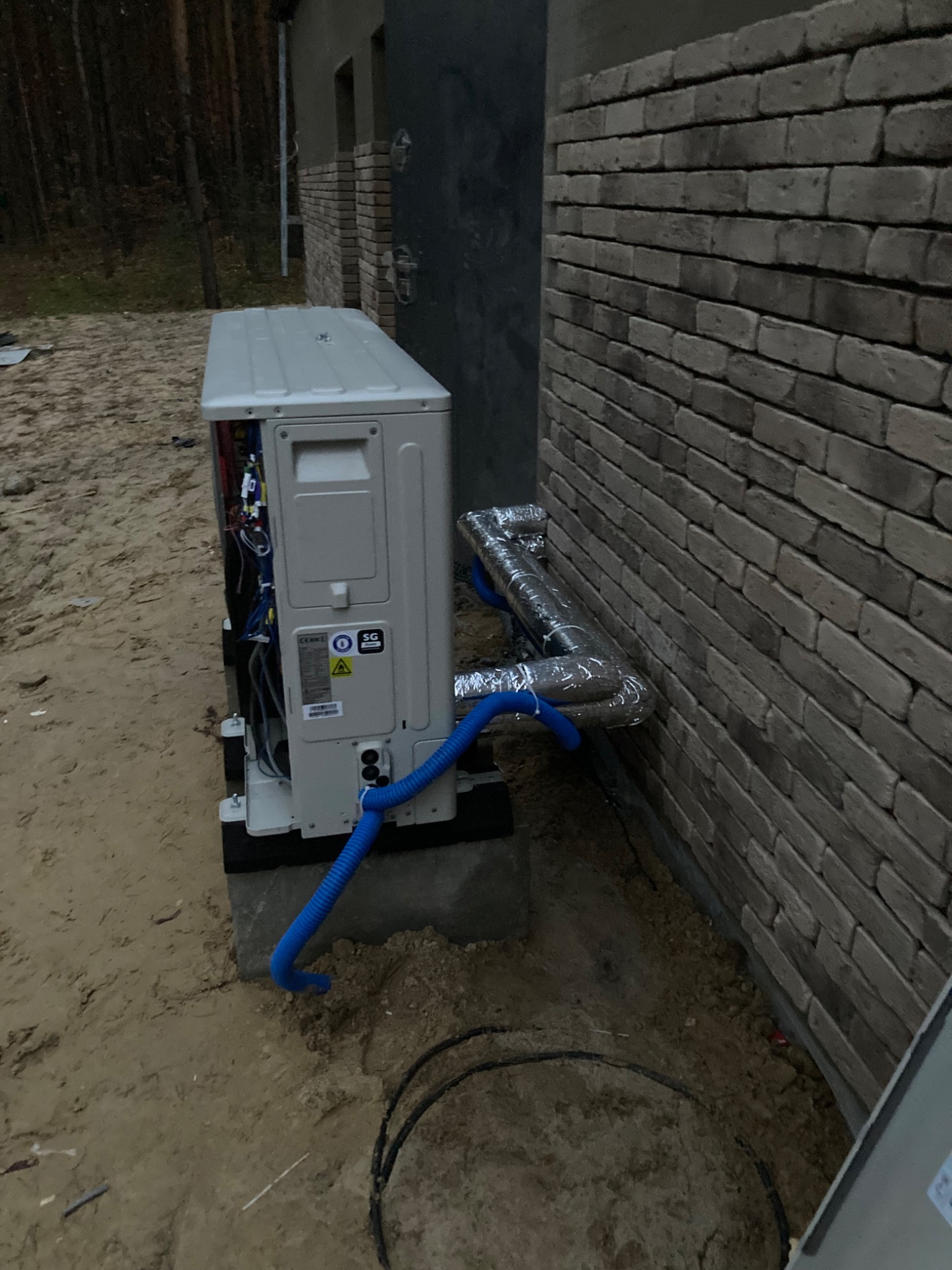 Zewnętrzna jednostka pompy ciepła zamontowana przy ceglanej ścianie budynku, z widocznymi niebieskimi rurami i zaizolowanymi przewodami, w surowym otoczeniu placu budowy.