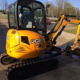 Żółta minikoparka JCB 8025 na gąsienicach, zaparkowana na betonowym podłożu, z widoczną kabiną i łyżką, w tle budynki mieszkalne i zieleń.