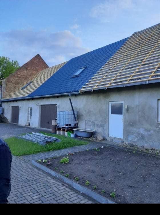 Częściowo pokryty niebieską dachówką dach budynku z widocznym oknem dachowym i drewnianą konstrukcją pod dalszą część pokrycia, na tle szarej elewacji i fragmentu ceglanego dachu sąsiedniego budynku.