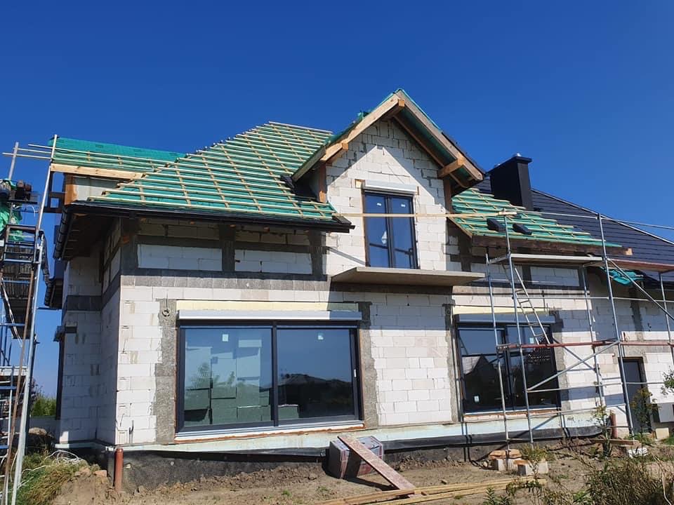 Budowa domu jednorodzinnego w trakcie, widoczne rusztowania, ściany z betonu komórkowego, częściowo ułożona dachówka w kolorze czarnym oraz drewniane elementy konstrukcji dachu przygotowane...