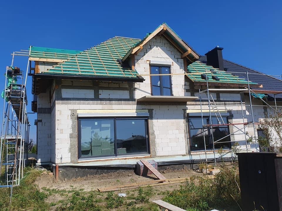 Budowa domu jednorodzinnego: widoczne rusztowania, częściowo położone deskowanie dachu z zieloną membraną, oraz fragment gotowego dachu z czarną dachówką.