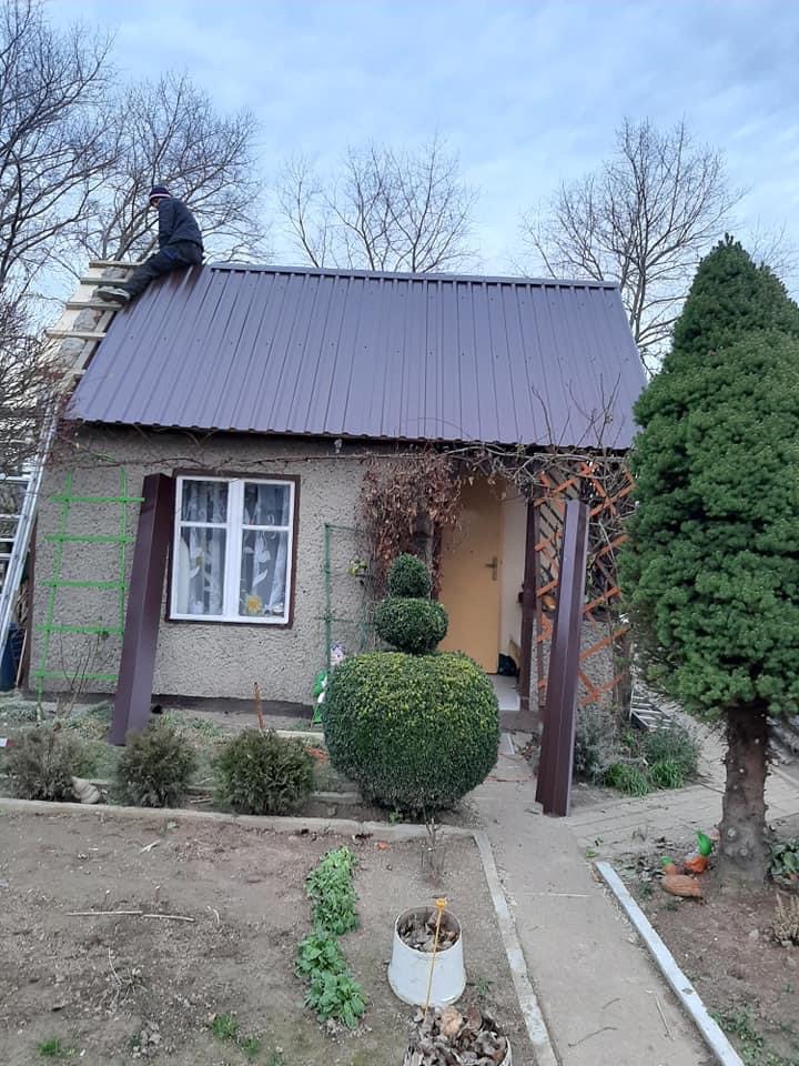 Praca na dachu niewielkiego domu z brązowym metalowym pokryciem, widoczny dekarz na drabinie, otoczenie domu z ogrodem i drzewami bez liści.