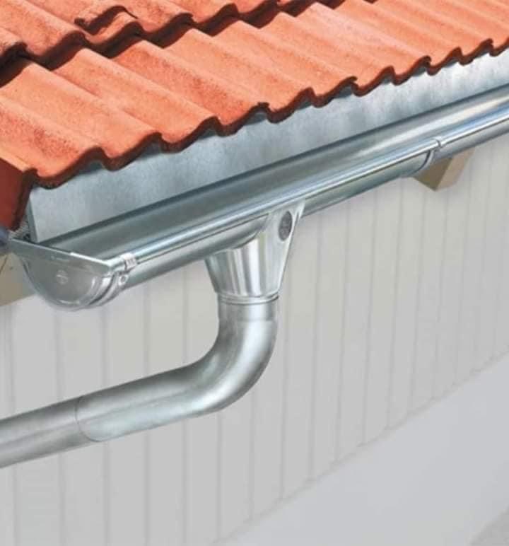 Nowa, błyszcząca, metalowa rynna zamontowana pod czerwonym dachem z dachówki ceramicznej, widoczna na tle białej elewacji budynku.