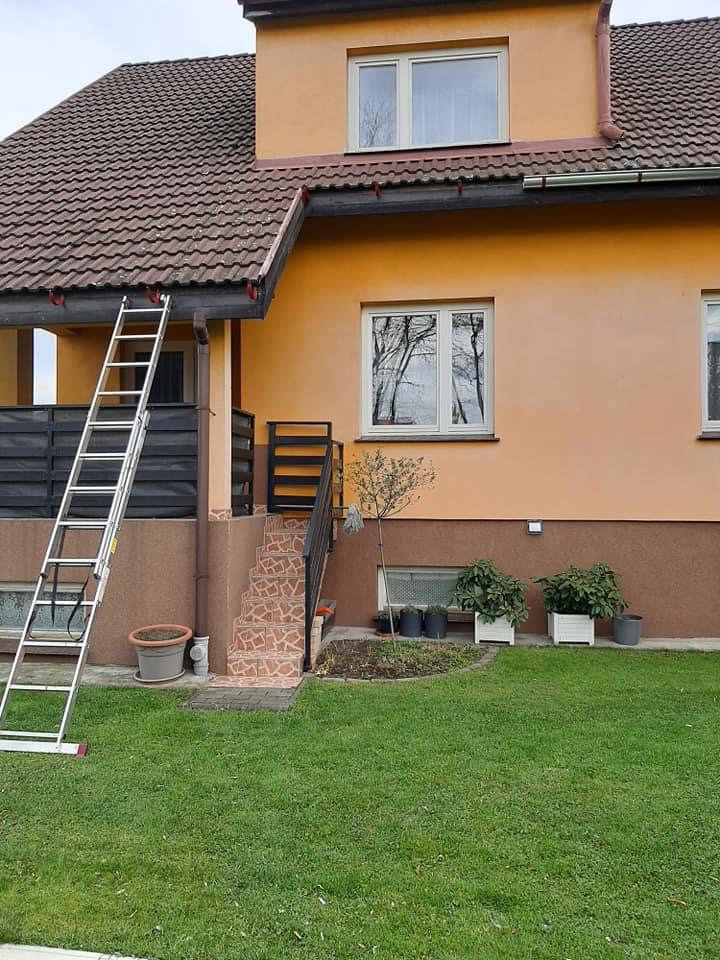 Dwupiętrowy dom z brązowym dachem i pomarańczową elewacją, drabina oparta o rynnę, widok z ogrodu.