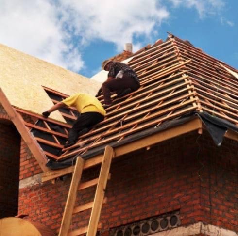 Dwóch dekarzy pracuje na drewnianej konstrukcji dachu z deskowaniem OSB, widoczna drabina oparta o ceglany budynek.