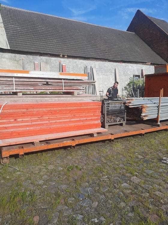 Przygotowane drewniane deski i metalowe rynny na platformie transportowej przed budynkiem z ciemnym dachem, dekarz w tle.