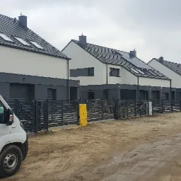 Szereg nowych domów jednorodzinnych z szarą stolarką okienną i ciemnymi dachami, widok z ulicy w trakcie prac wykończeniowych, z zaparkowanym białym busem.