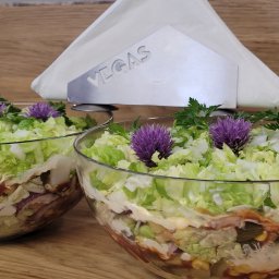 Vegas Grzegorz Wiśniowski - Dwie szklane miski z warstwową sałatką, udekorowane fioletowymi kwiatami i zieloną pietruszką. W tle podajnik na serwetki z napisem 'VEGAS'.