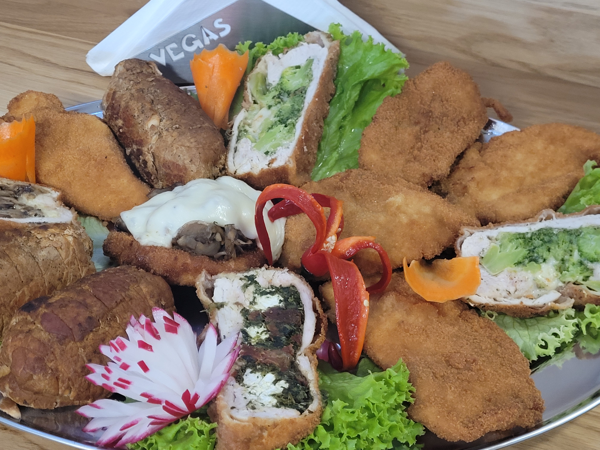 Dekoracyjna srebrna patera z roladkami drobiowymi, mięsem i warzywami, w tym rzodkiewką w kształcie kwiatu. Widoczny napis Vegas na serwetniku.