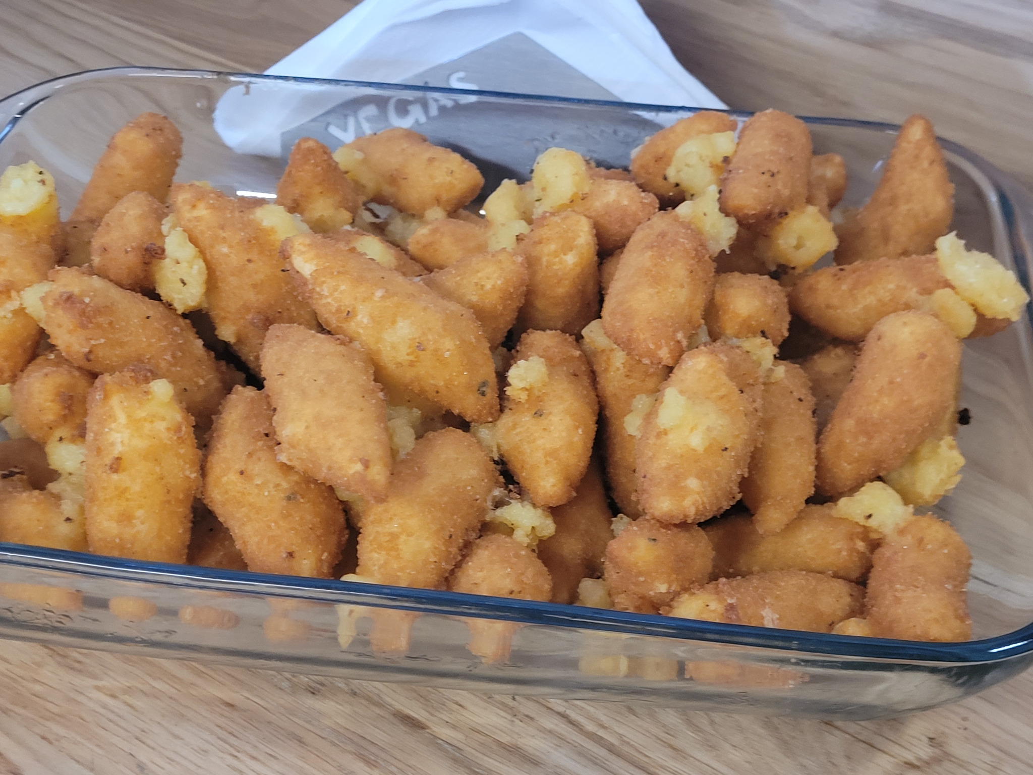 Złociste krokiety ziemniaczane w szklanym naczyniu, widoczne z bliska, na drewnianym blacie. Część krokietów z widocznym farszem ziemniaczanym.