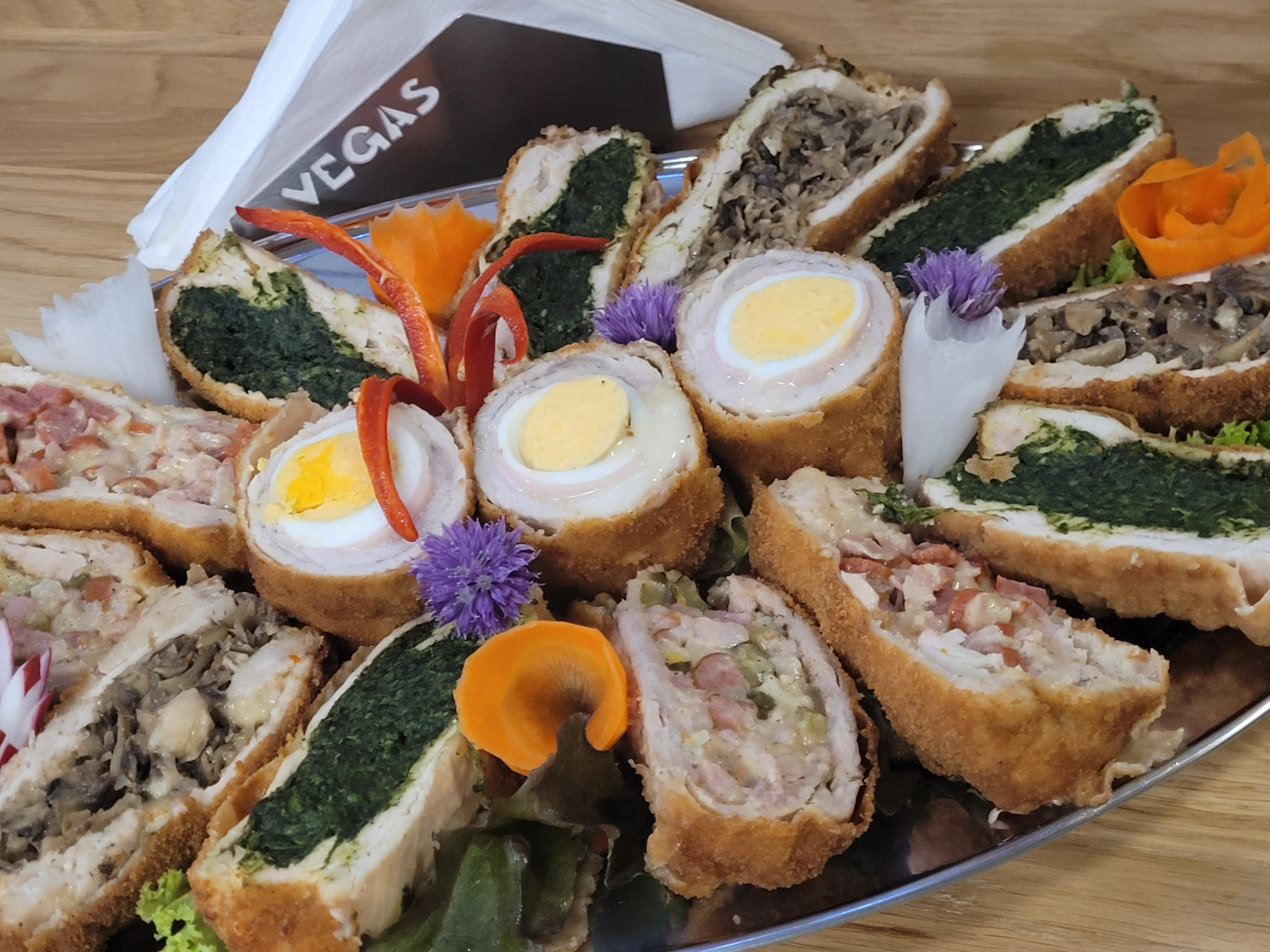 Elegancki talerz z roladkami z różnymi nadzieniami (szpinak, pieczarki, jajko), dekorowany warzywami i kwiatami, z serwetkami 'Vegas' w tle.