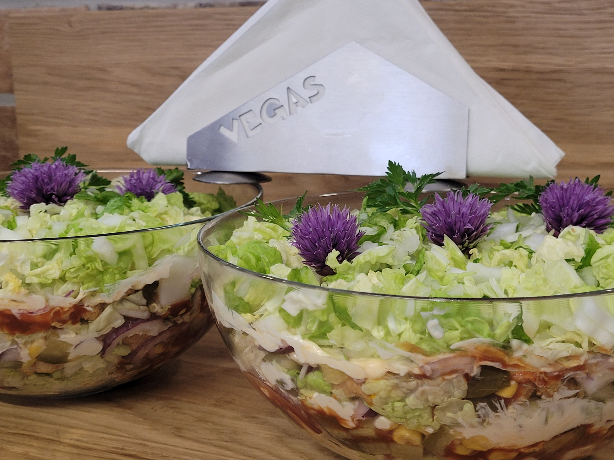 Dwie szklane miski z warstwową sałatką, udekorowane fioletowymi kwiatami i zieloną pietruszką. W tle podajnik na serwetki z napisem 'VEGAS'.