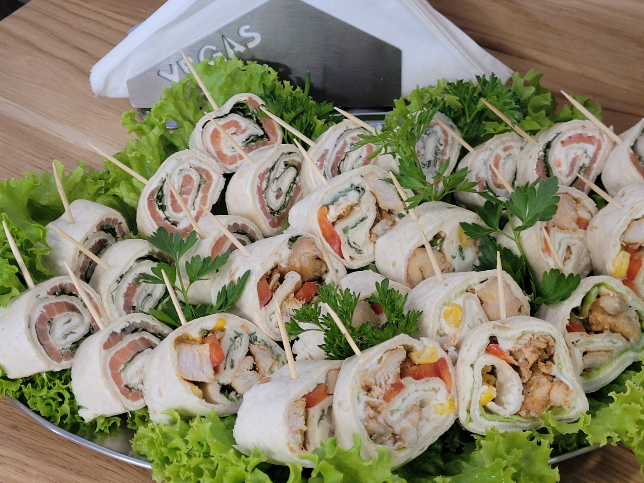 Elegancko podane roladki tortilli z różnymi nadzieniami (łosoś, kurczak) na srebrnej tacy, udekorowane sałatą i ziołami. Szpilki utrzymują zwinięte kawałki.