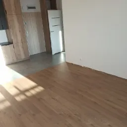 Nowe panele podłogowe w jasnym odcieniu drewna, ułożone w pokoju z białymi ścianami i fragmentem zabudowy kuchennej oraz dwiema parami drzwi.