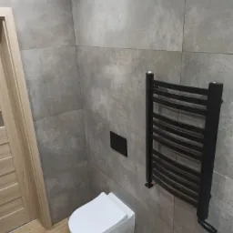 Nowoczesna łazienka z szarymi płytkami imitującymi kamień, wisząca toaleta i czarny grzejnik drabinkowy.