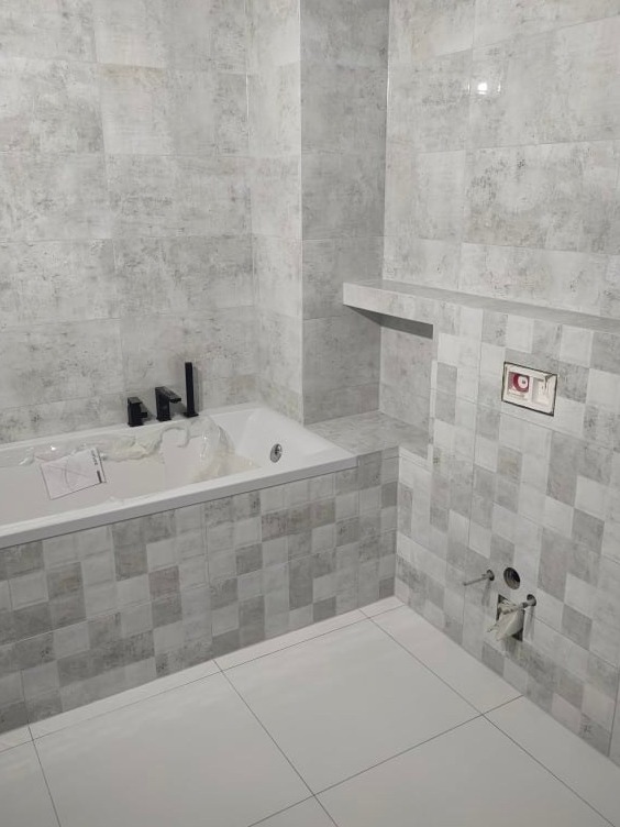 Wykończona łazienka z wanną obłożoną mozaiką w odcieniach szarości, ściany wyłożone płytkami imitującymi beton, czarna armatura, biała podłoga z płytek.