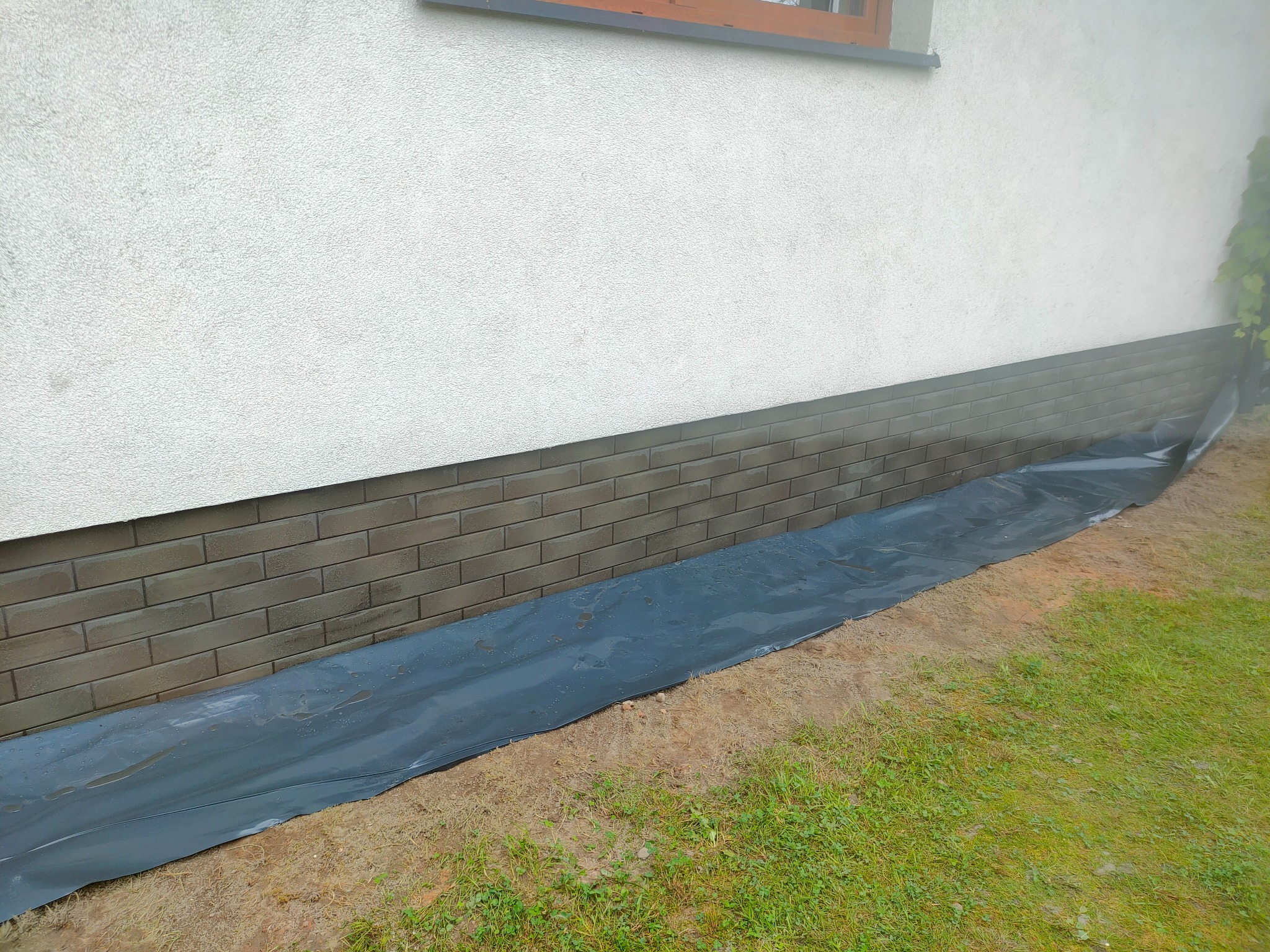 Szara ceglana podmurówka budynku, zabezpieczona czarną folią ochronną na trawniku.