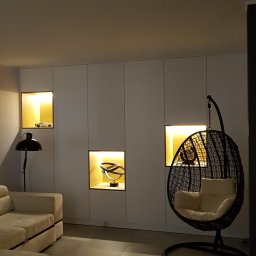 MZW - Białe szafy wnękowe z podświetlanymi niszami, w których znajdują się dekoracje: model samochodu, abstrakcyjna rzeźba i plecione krzesło wiszące z poduszką.