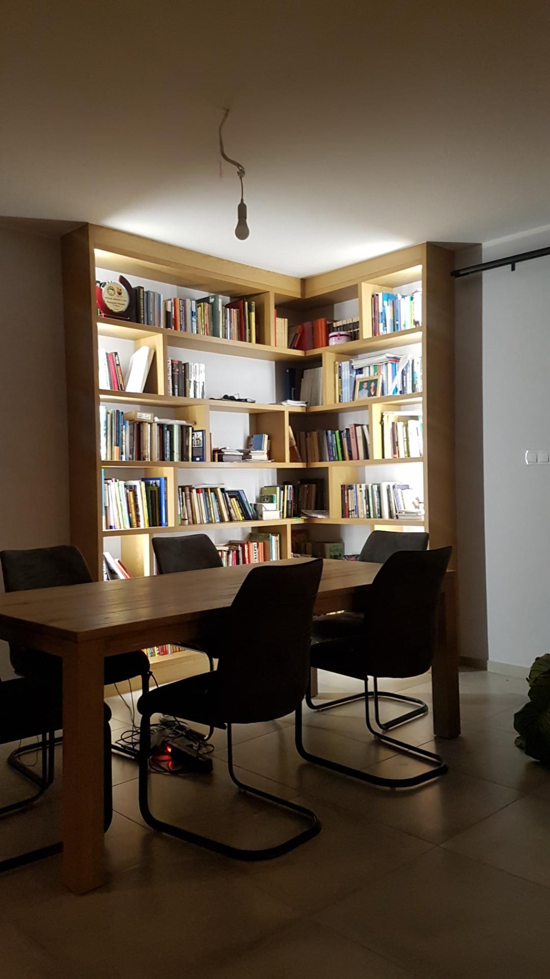Drewniana biblioteczka narożna z podświetleniem, wypełniona książkami, widoczna zza stołu jadalnego z krzesłami. Na suficie zwisa żarówka na kablu.