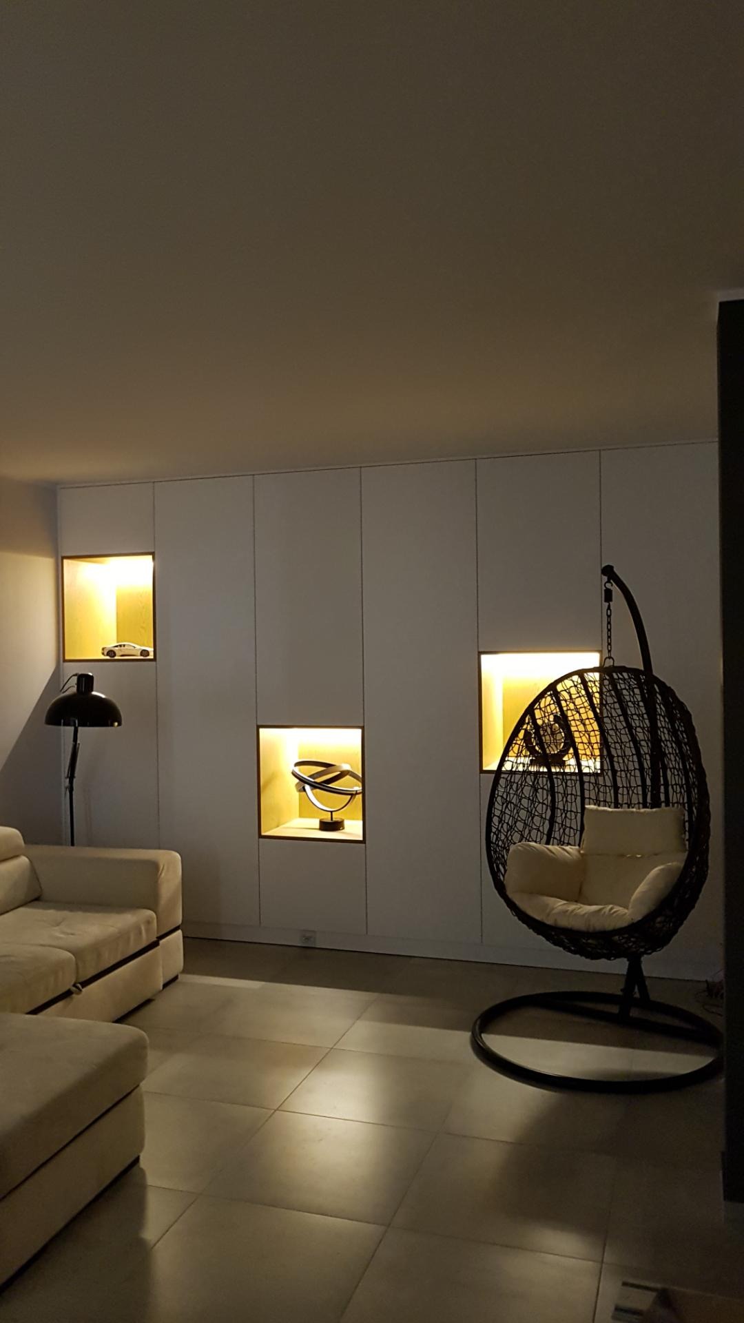 Białe szafy wnękowe z podświetlanymi niszami, w których znajdują się dekoracje: model samochodu, abstrakcyjna rzeźba i plecione krzesło wiszące z poduszką.