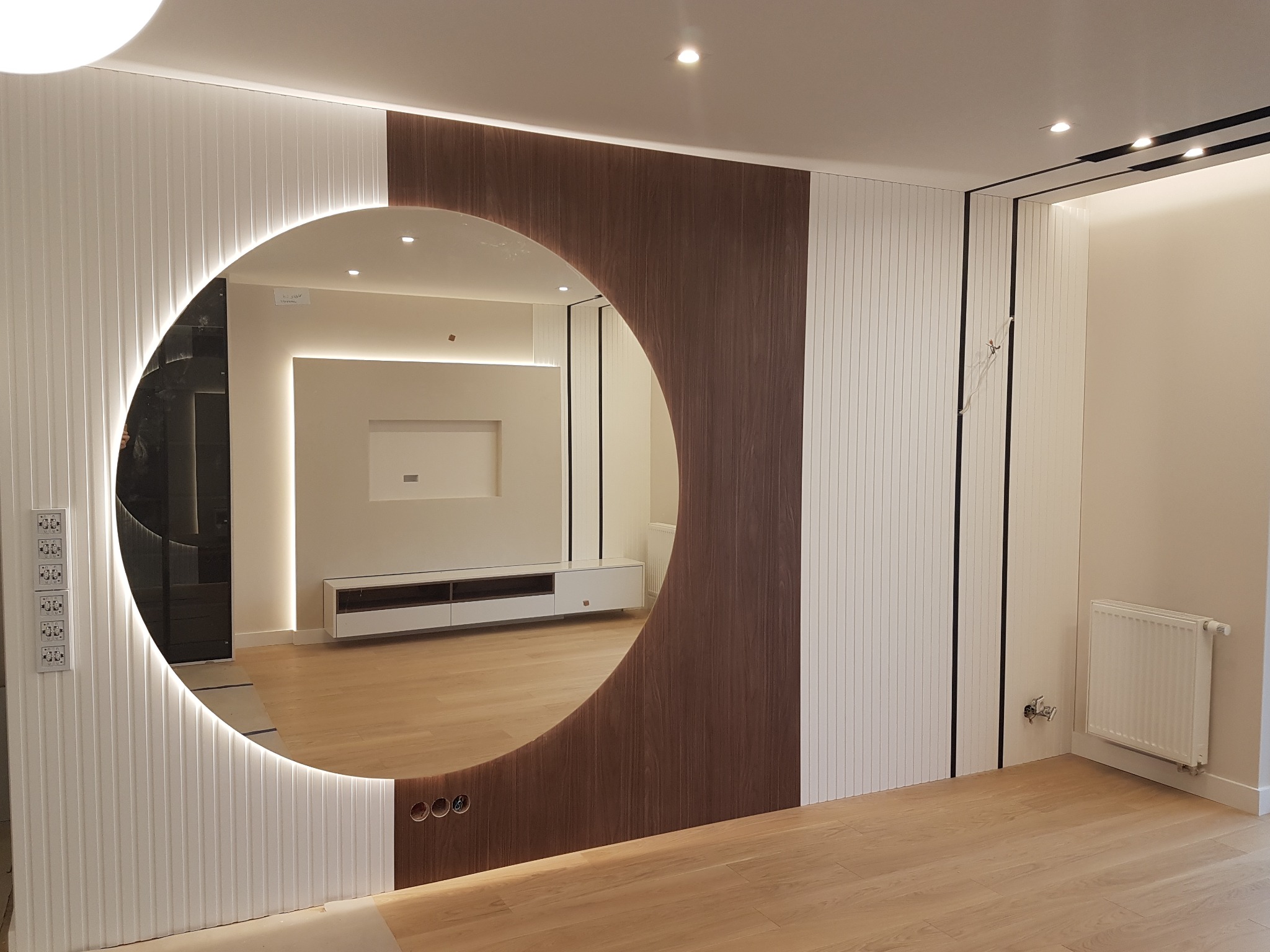 Nowoczesna aranżacja wnętrza z okrągłym lustrem z podświetleniem LED, obudowanym drewnianą i białą panelową ścianą, odbijającym minimalistyczny salon z szafką RTV i oświetleniem punktowym