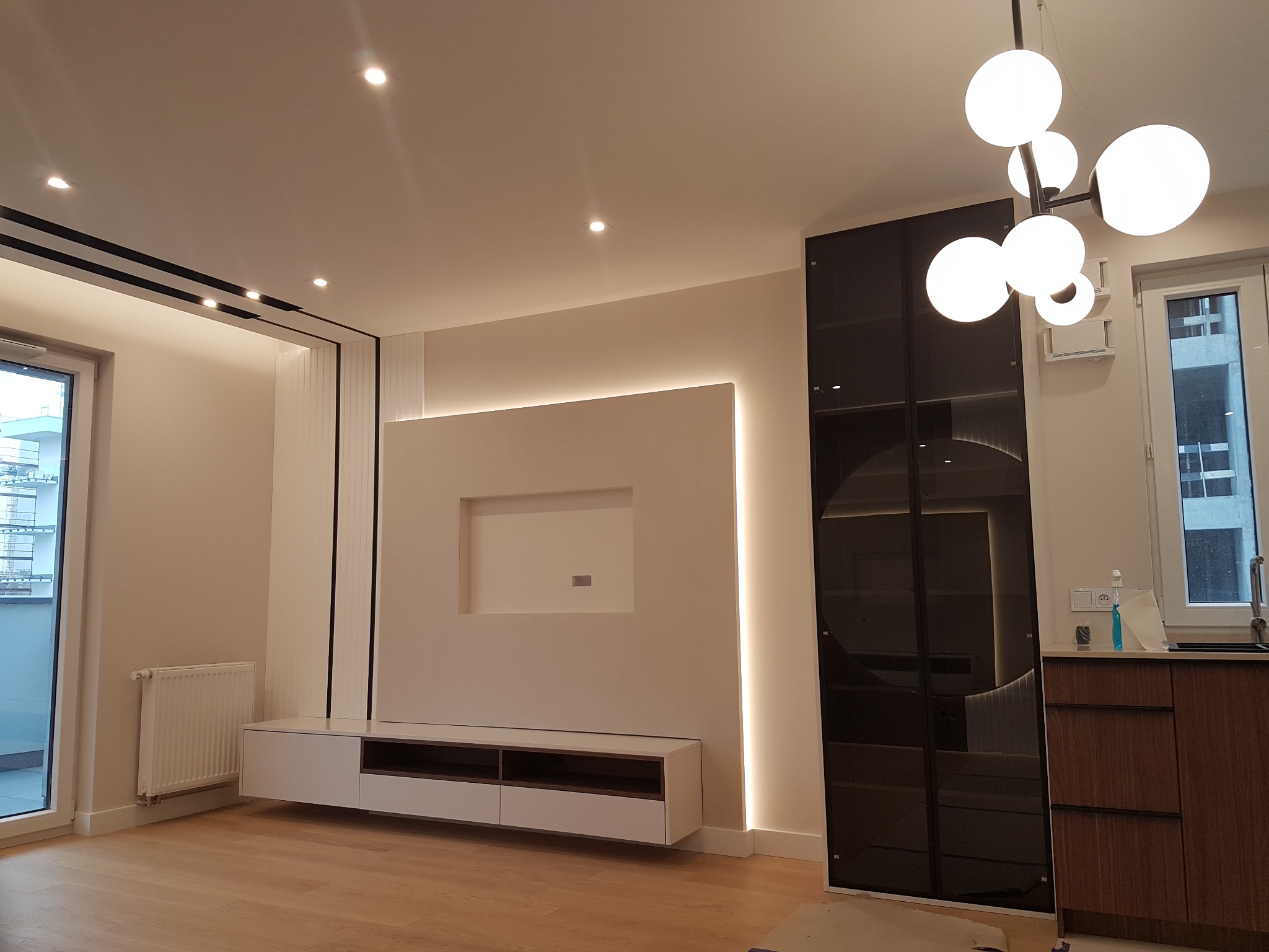 Nowoczesny salon z zabudową meblową: biała szafka RTV z wnękami, podświetlana ściana z wnęką na telewizor, wysoka szafa z czarnymi, szklanymi drzwiami i drewniane blaty. Całość oświetlona...