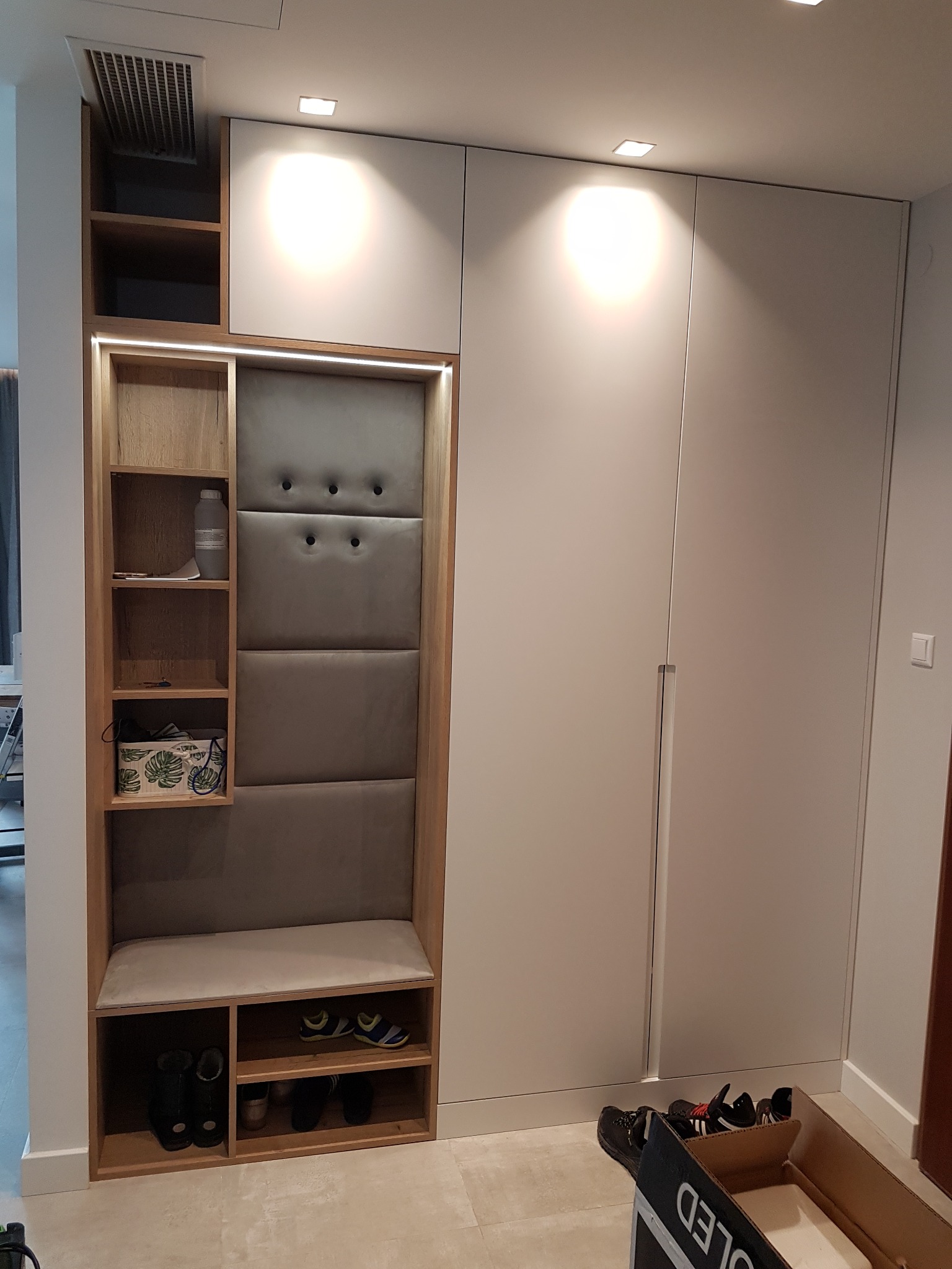 Wnętrze przedpokoju z zabudową: biała szafa z drewnianym regałem, siedziskiem i tapicerowanym panelem ściennym; na półkach dekoracje i buty.