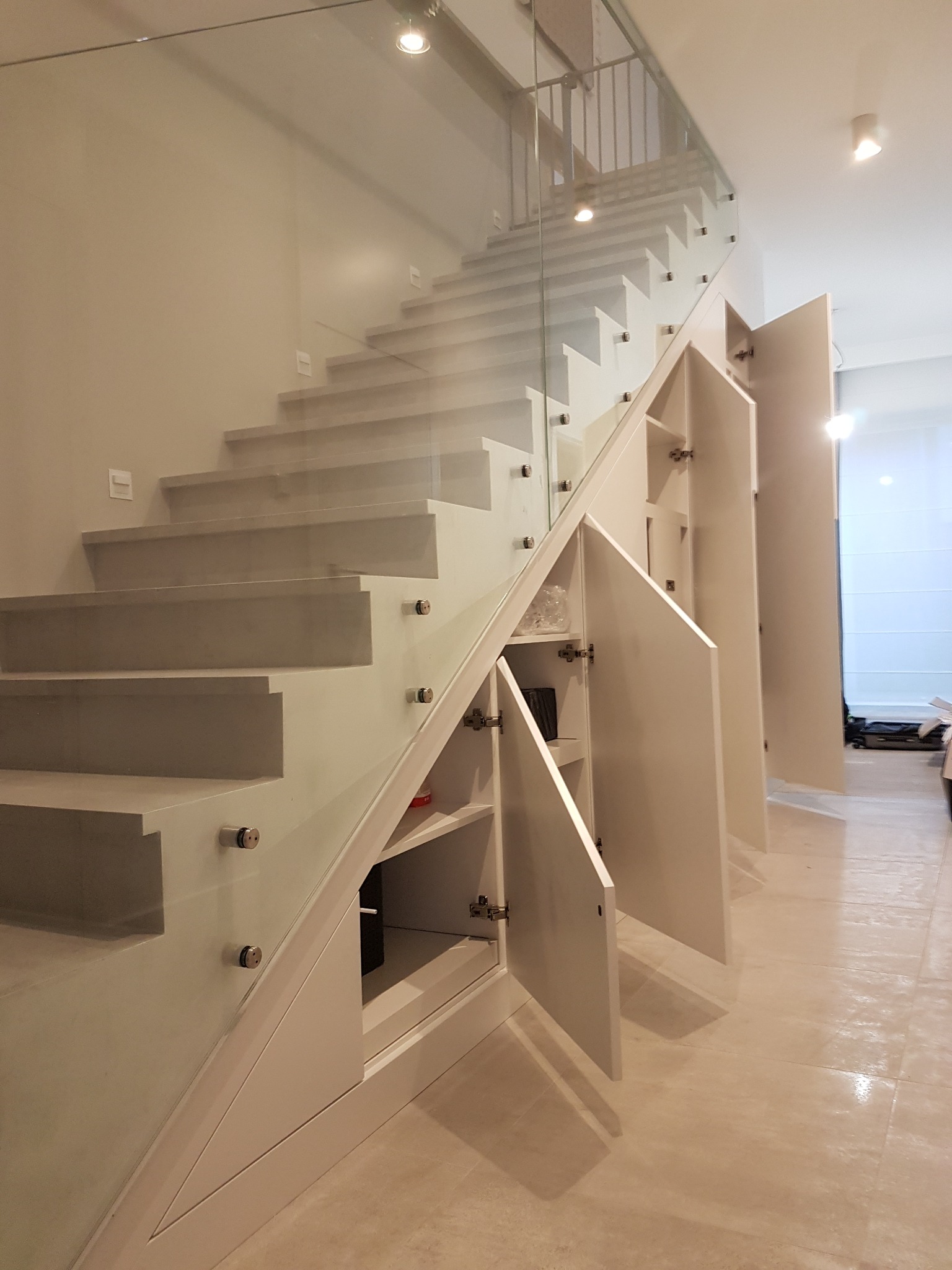Białe schody z szklaną balustradą i szafkami w przestrzeni pod schodami z uchylonymi drzwiczkami.