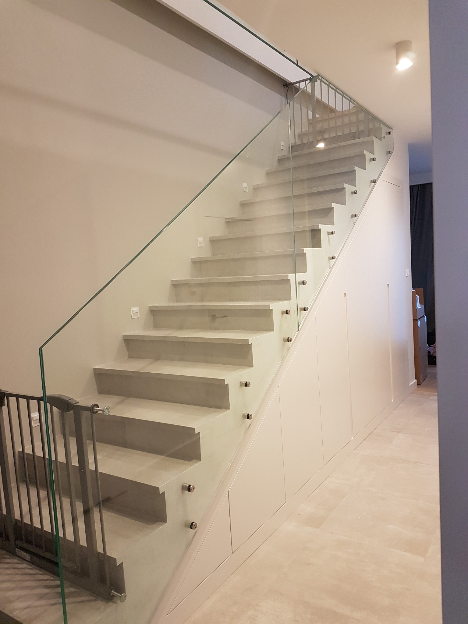Schody wewnętrzne z szarymi stopniami i szklaną balustradą mocowaną na metalowych uchwytach, zabudowa szafy pod schodami z białymi frontami.