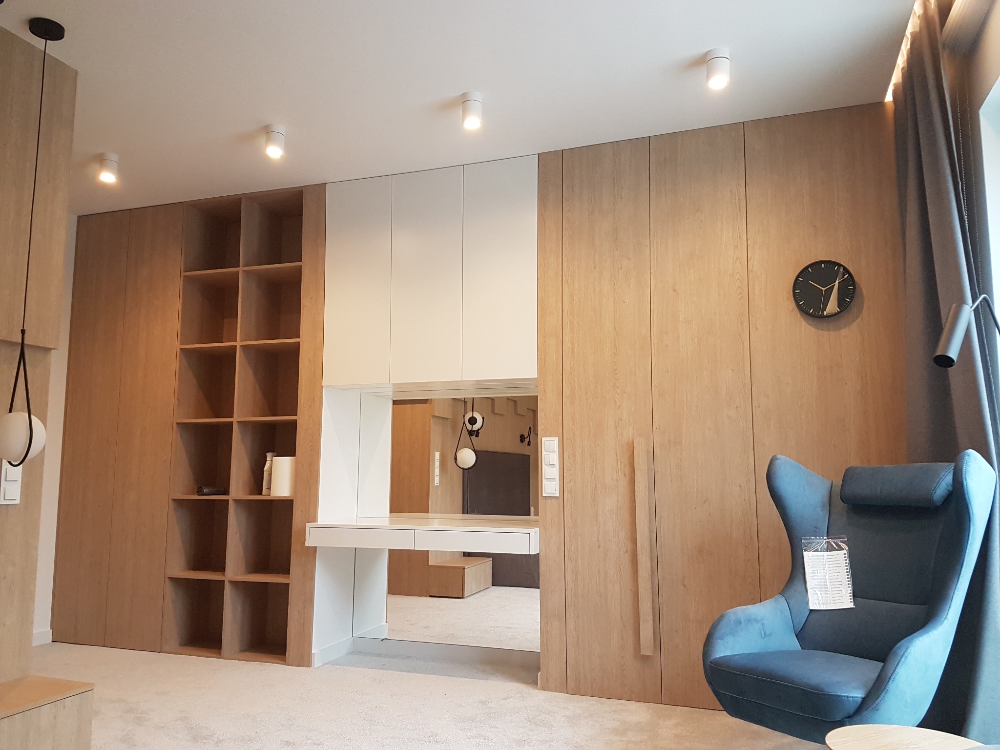Zabudowa ściany z jasnego drewna z półkami, szafkami i biurkiem z lustrem, uzupełniona o niebieski fotel z wysokim oparciem i czarny zegar na ścianie.