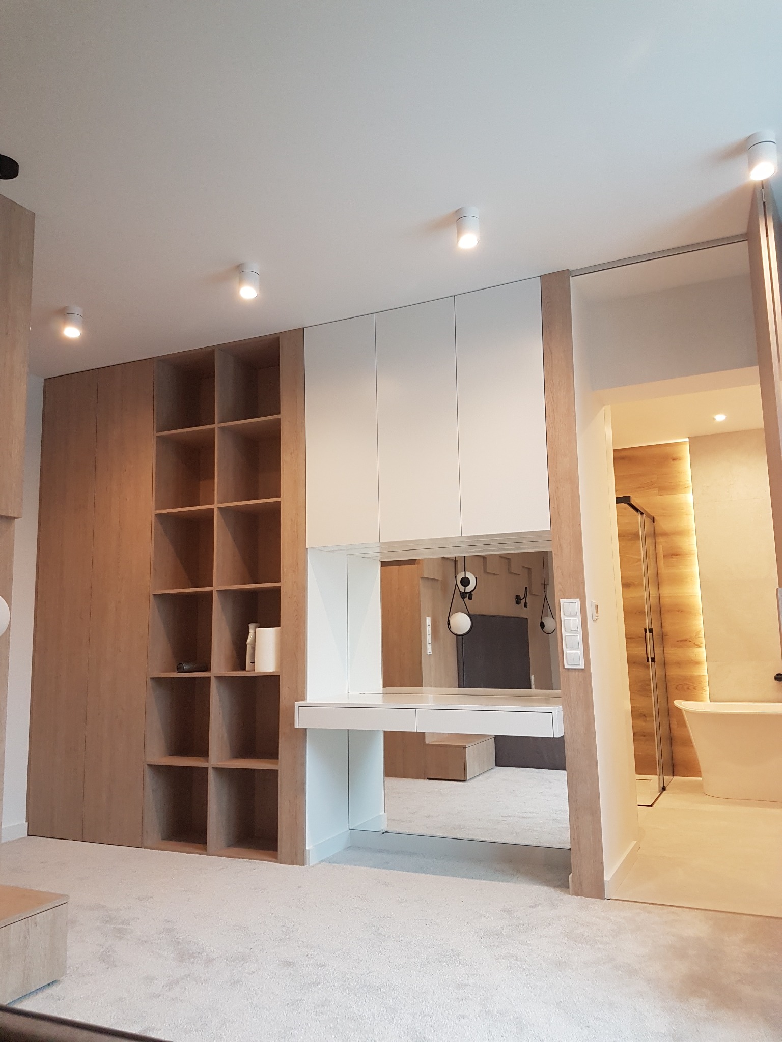 Nowoczesna zabudowa garderoby z drewnianymi elementami i białymi szafkami wiszącymi, połączona z toaletką i lustrem, z widokiem na łazienkę z wanną wolnostojącą.