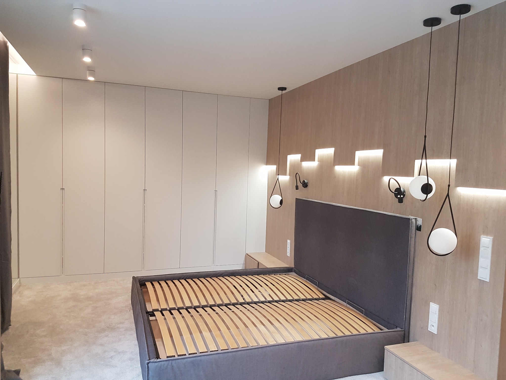 Nowoczesna sypialnia z łóżkiem bez materaca, drewnianym stelażem i tapicerowanym zagłówkiem, oświetleniem LED na ścianie i minimalistycznymi lampami wiszącymi, oraz szafą w zabudowie w tle.