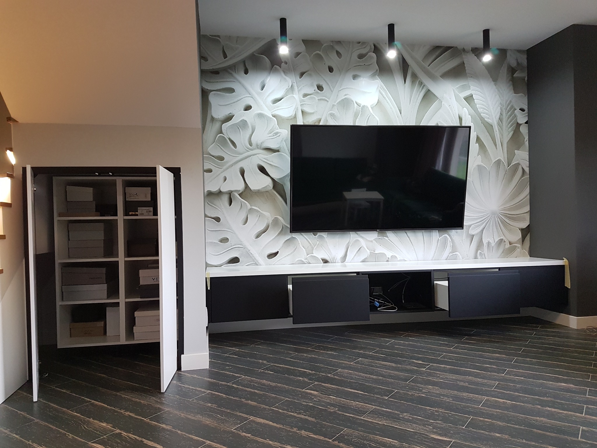 Nowoczesny salon z szafką RTV w kolorze czarnym, białą szafą na buty z otwartymi drzwiami i dekoracyjną ścianą z motywem liści, oświetloną trzema reflektorami.