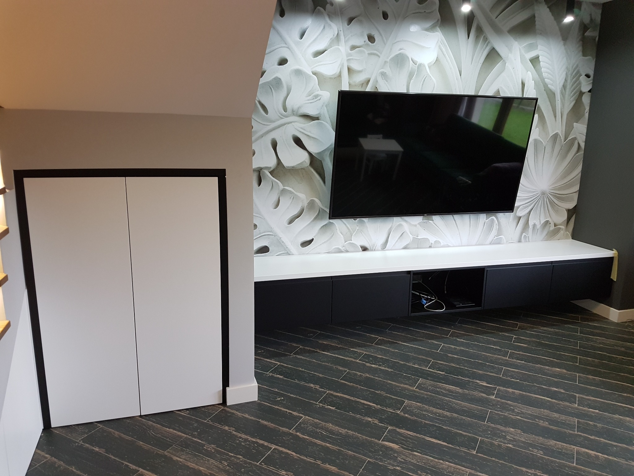 Nowoczesny salon z białą szafą wnękową z czarnymi obramowaniami, podwieszaną szafką RTV w kolorze czarno-białym oraz ścianą z dekoracyjną tapetą 3D z motywem liści.
